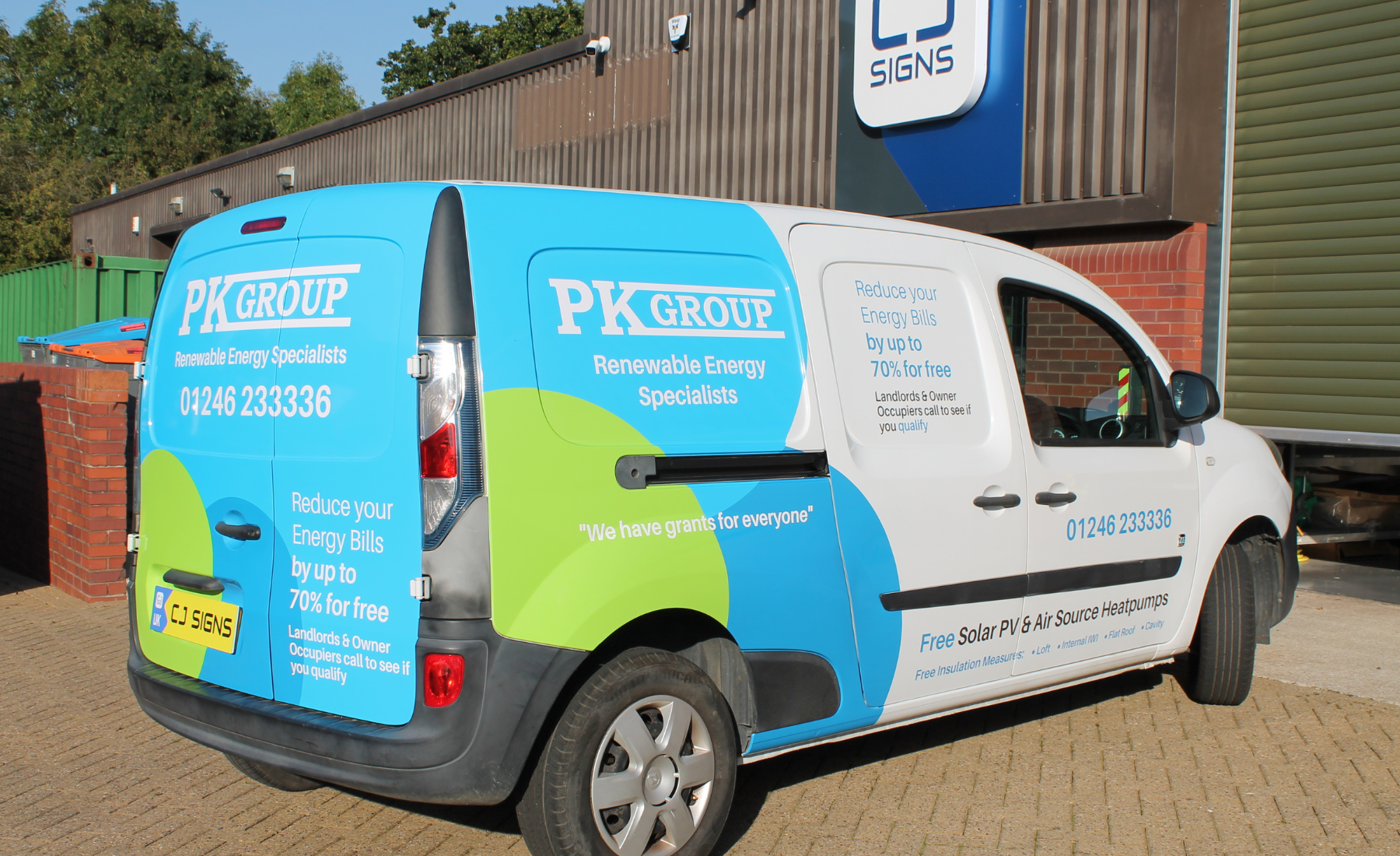A PK Group company van with branded vehicle wrap promoting renewable energy services. The van displays text including 'Reduce your Energy Bills by up to 70% for free,' 'We have grants for everyone,' and contact details including the phone number 01246 233336. The van is parked outside CJ Signs, the signage company responsible for the wrap.